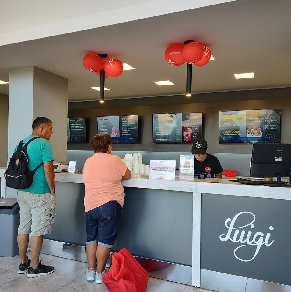 Mostrador de un local Luigi con clientes siendo atendidos frente a las pantallas de menú y el logo de la marca.